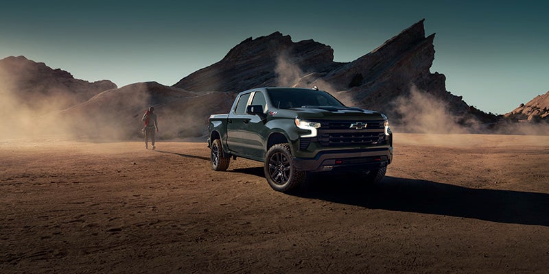 Gray Chevrolet Silverado on a dirt road with mountains and a man in the background